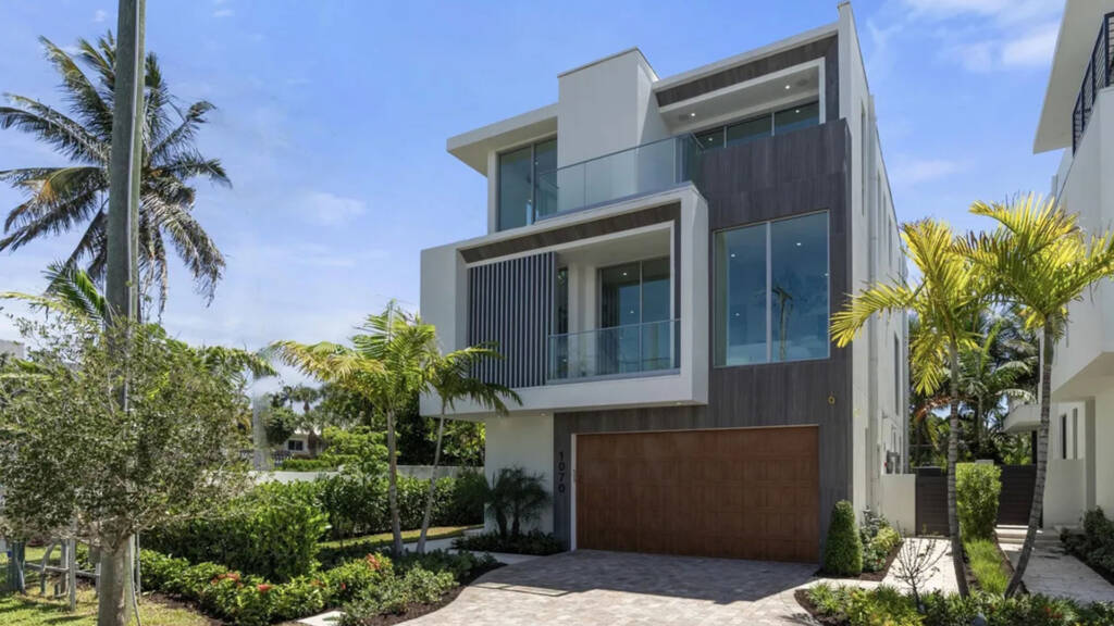 An exterior view of a modern three-story luxury home at Ocean Delray Estates in Delray Beach, FL. The residence features contemporary coastal architecture with wood-textured siding, glass-railing balconies, oversized windows, and a rich wood-finish garage door. The property is professionally landscaped with palm trees and lush greenery under a clear blue sky.