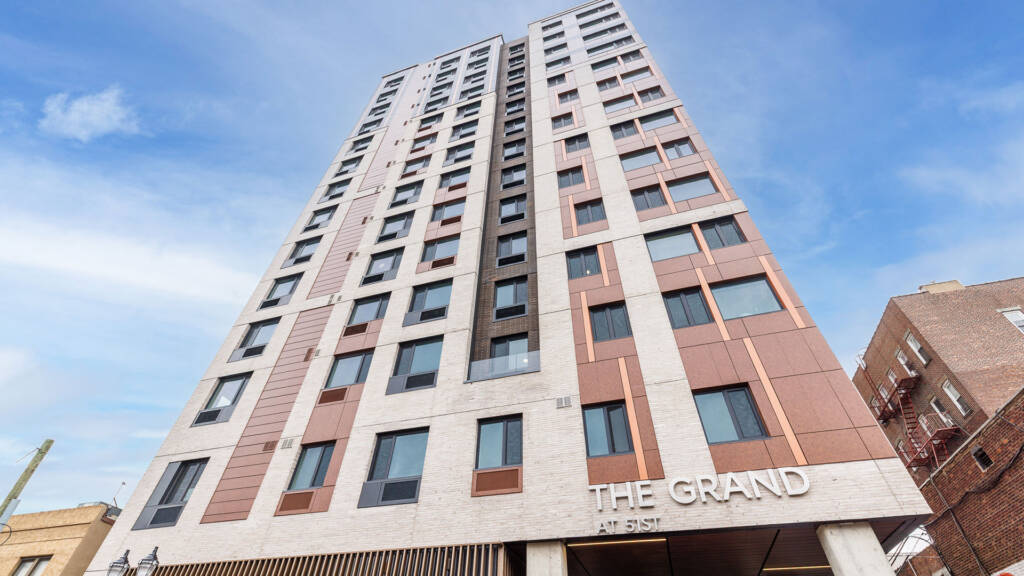 A low-angle exterior view of The Grand at 51st, a modern high-rise apartment building in West New York, NJ featuring a white brick and copper-toned paneled facade.